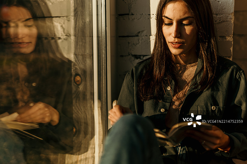 年轻女性在金色阳光下靠窗阅读一本书图片素材