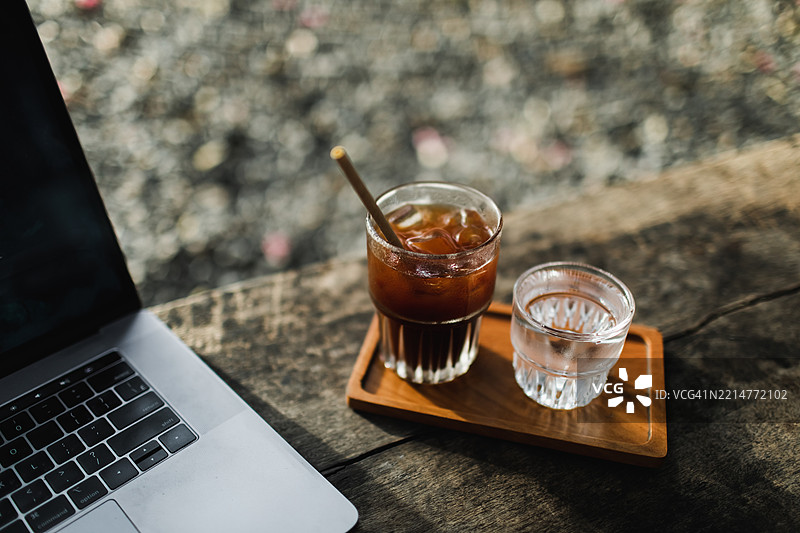 户外咖啡馆木桌上的清爽冰黑咖啡和笔记本电脑。自由职业者工作场所图片素材