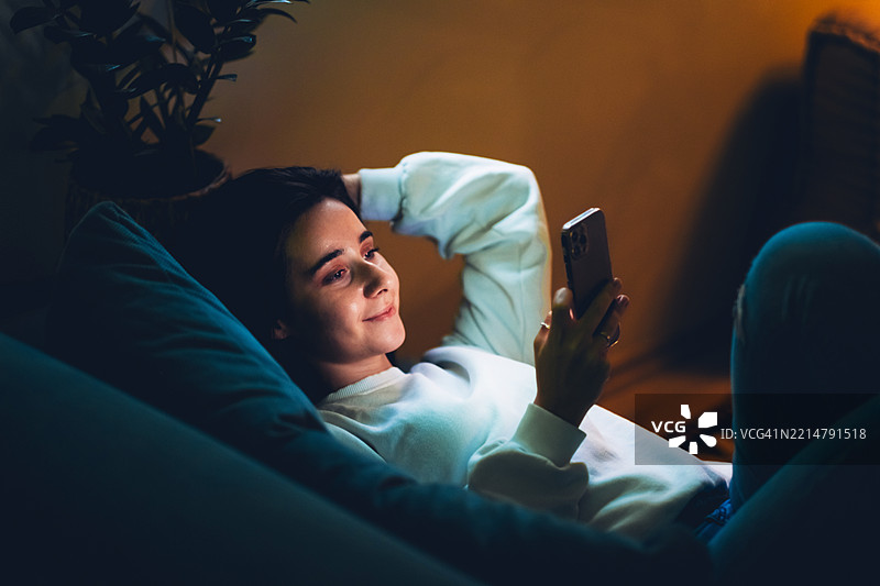在家中享受轻松的晚上，舒适温馨的客厅氛围。年轻的黑发女性躺在沙发上使用智能手机。温馨的室内灯光，休闲的服装，轻松的心情。图片素材