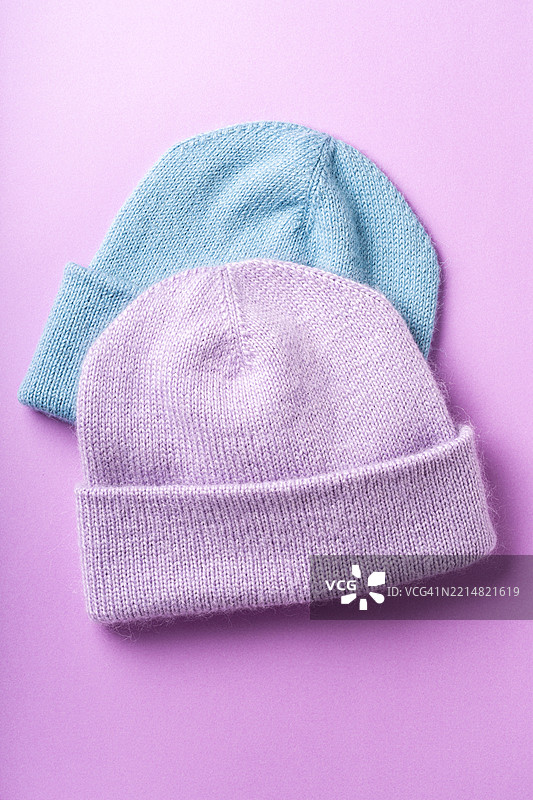 两顶舒适的针织毛帽，柔和的粉彩色，一顶浅蓝色，一顶薰衣草色，静静地放在鲜艳的紫色背景上，展现冬季时尚的温暖与风格。图片素材