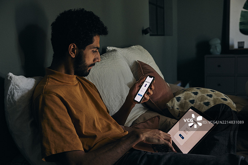 男子在卧室里坐着，使用智能手机输入笔记本电脑的认证密码图片素材