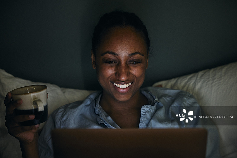微笑的女性在夜晚的卧室里一边使用笔记本电脑一边拿着杯子图片素材