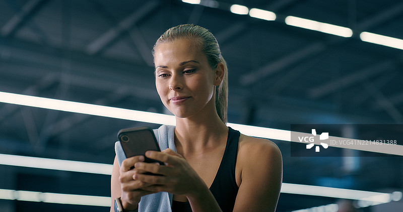 健身房里的女性，使用智能手机聊天，休息时查看训练的运动数据和移动应用。此人微笑着滑动手机，联系或社交媒体，进行健身锻炼。图片素材