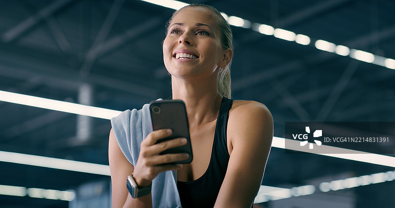 女性在健身房愉快地使用手机，休息并查看训练的运动数据。此人使用智能手机与聊天、联系或社交媒体互动，享受健身俱乐部的锻炼时光。图片素材