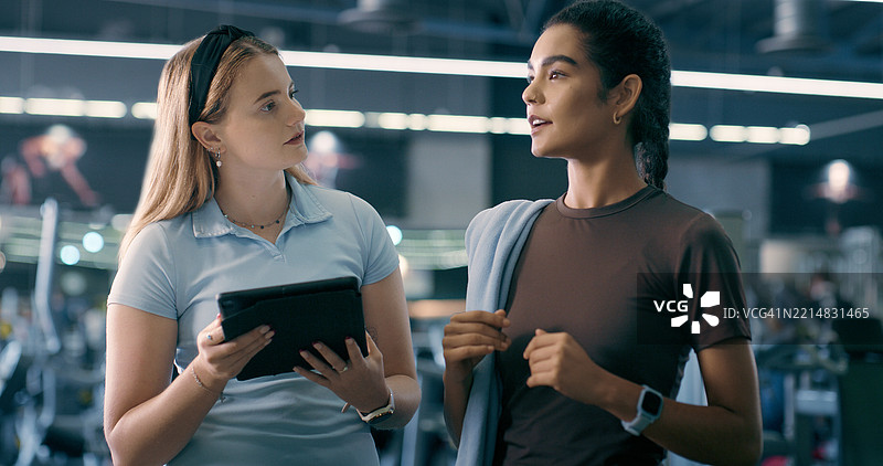 健身房内的女性私人教练与平板电脑进行健身计划讨论，支持身体目标或锻炼交流。教练、科技与健身、清单或健康发展的讨论。图片素材