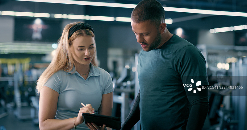 健身房内的女性私人教练与平板电脑进行锻炼计划讨论，支持身体目标或健身交流。教练、科技与健身、清单或健康发展的讨论。图片素材