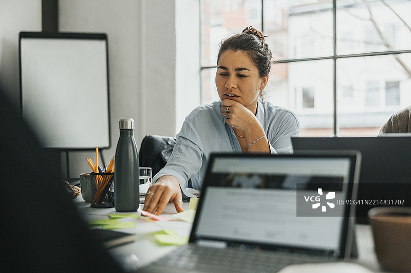 年轻女性商务专业人士坐在科技初创公司的会议室里，手托着下巴，阅读便签。图片素材