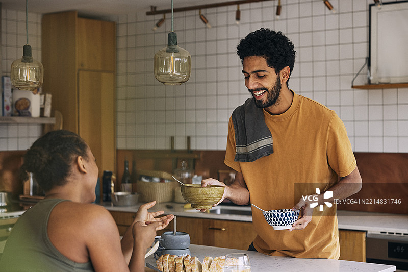 快乐的男人在家中将碗递给坐在桌旁的女友图片素材