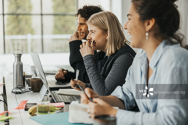 快乐的金发女性IT专业人士坐在初创公司办公室里，身边有同事们，使用着笔记本电脑。图片素材