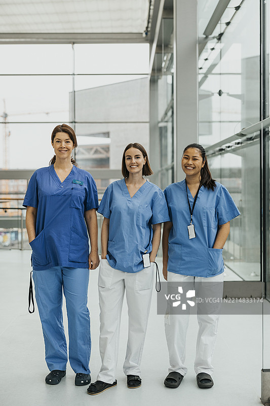 微笑的女性医疗专家并肩站在医院走廊，手插在口袋里图片素材