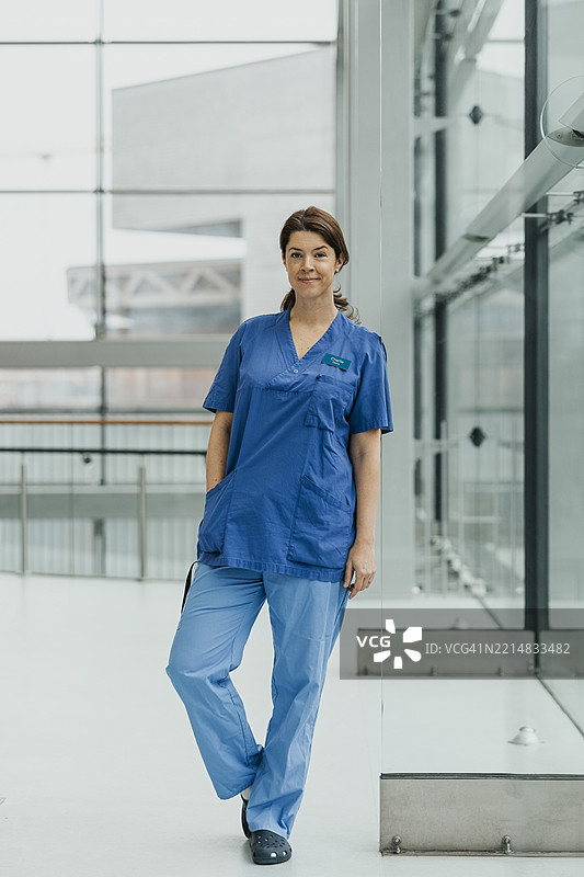 自信的女性医护人员穿着医疗制服，靠在医院的玻璃墙上，手插在口袋里。图片素材