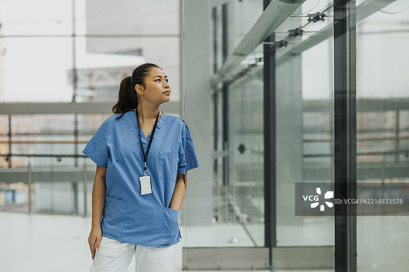 年轻女医生在医院走廊里站着，手插在口袋里，透过玻璃墙观察。图片素材