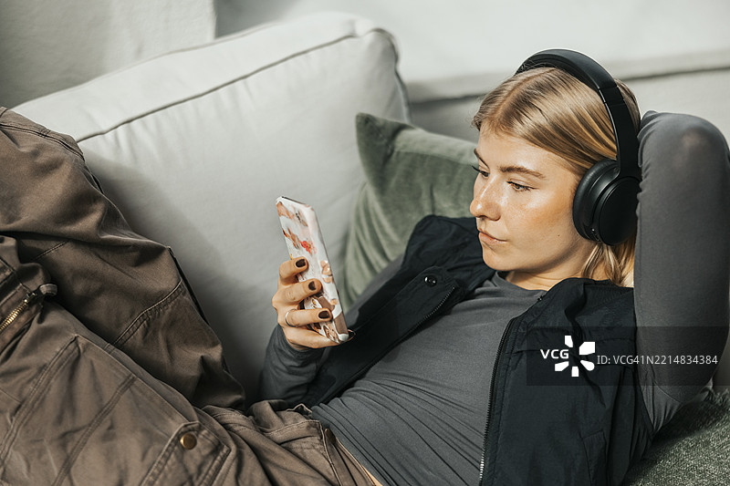 高角度视图的金发IT专业人士在办公室沙发上躺着，使用智能手机并佩戴无线耳机。图片素材