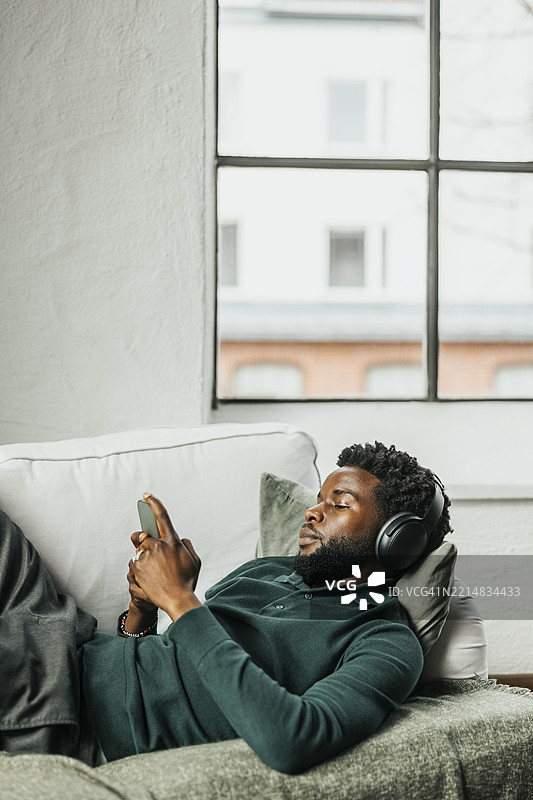 放松的商人躺在沙发上，戴着无线耳机，使用智能手机，身处办公室。图片素材