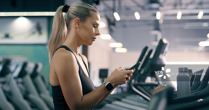 女性在健身房使用手机输入健身应用程序、锻炼计划或运动日程。活跃的女性用户使用手机跟踪健康俱乐部的表现或卡路里结果。图片素材