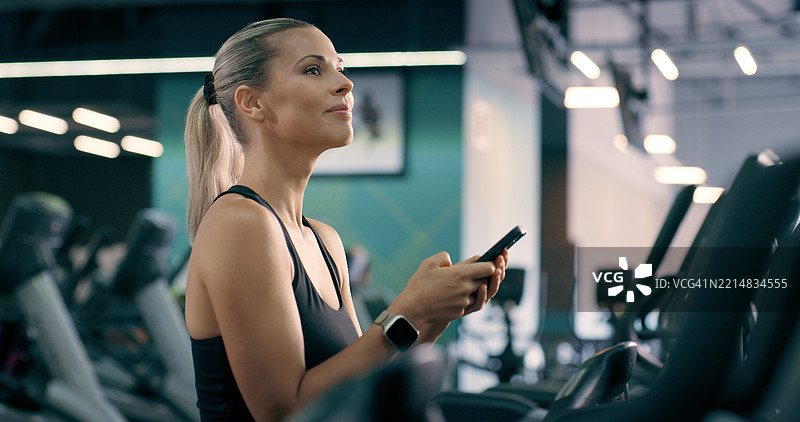 健身房里的女性，手持智能手机，思考社交媒体和数字应用以跟踪进展。健康、决策或在健康中心的人，使用手机进行锻炼，查看电子邮件并深思。图片素材