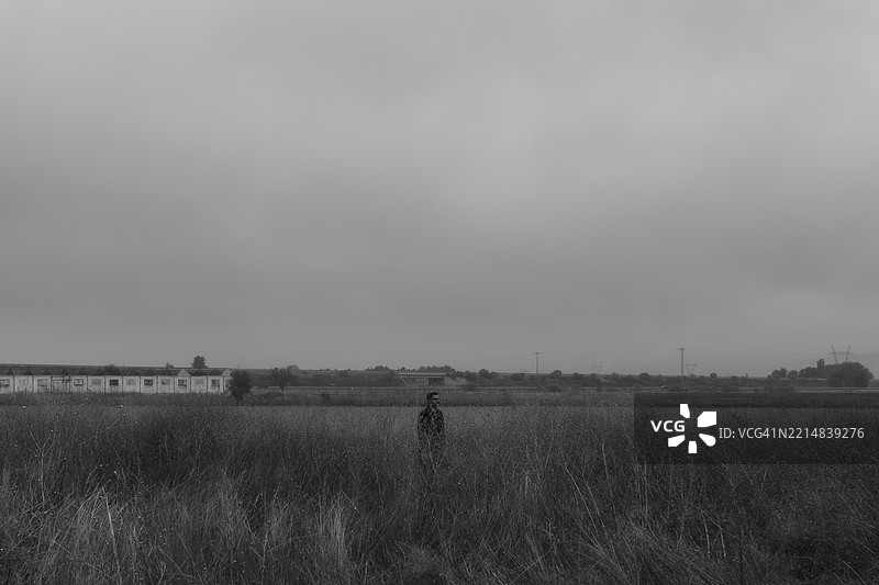 孤独的男人站在阴天的高草中，背景是废弃的建筑。图片素材