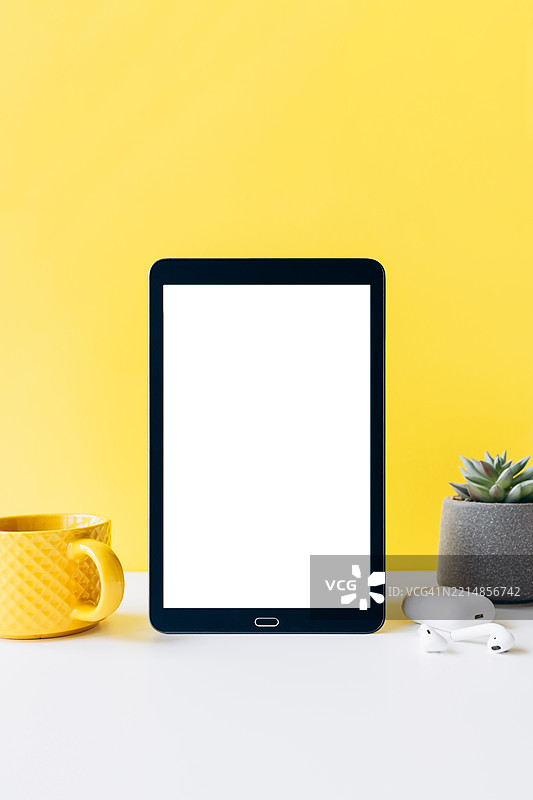 白色桌面上的数字平板电脑，屏幕空白，旁边有一个黄色咖啡杯、无线耳机和一盆植物——现代工作空间概念图片素材