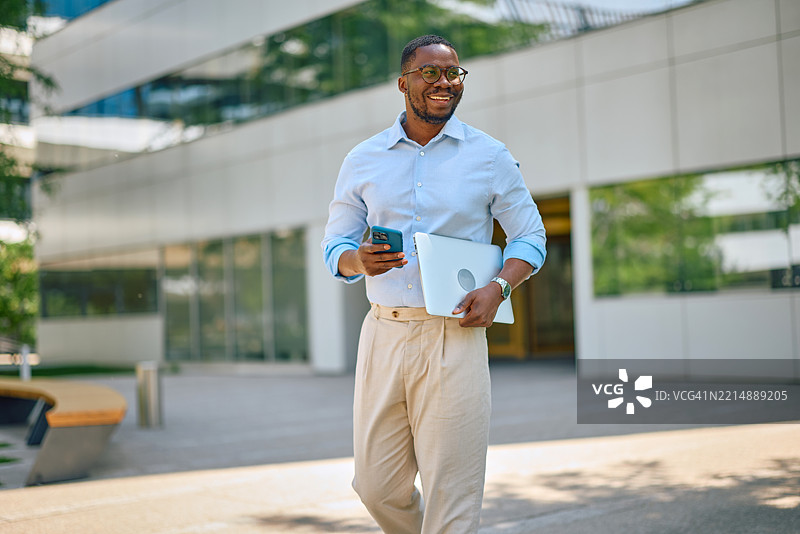 微笑的商人在办公大楼前走路，使用智能手机并手持笔记本电脑。图片素材