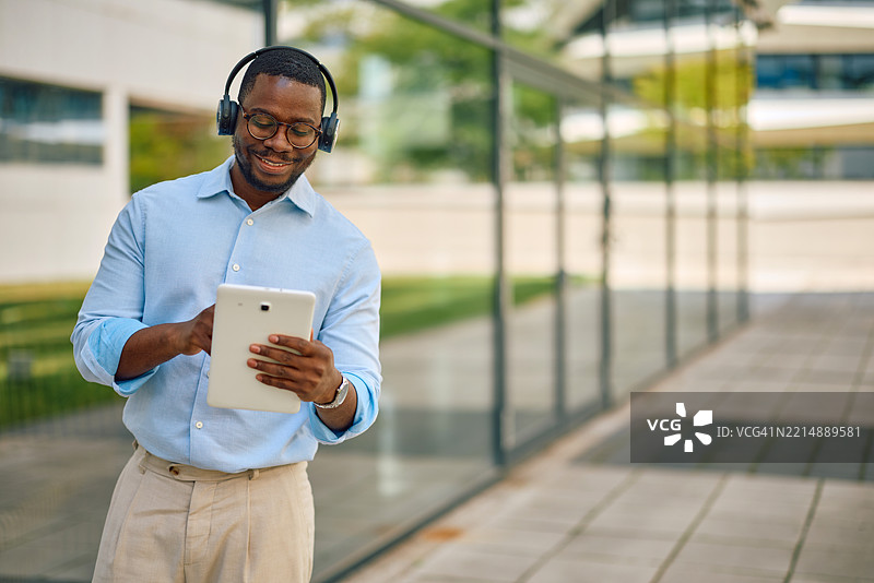 微笑的商人在户外使用平板电脑并戴着耳机听音乐图片素材