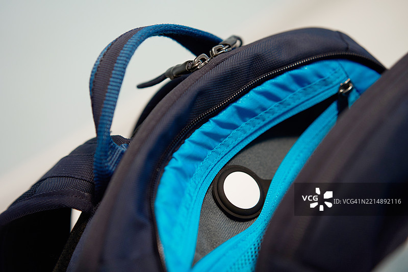 蓝色背包前隔层内隐藏的GPS追踪器AirTag，拉链打开，背景为浅色。图片素材