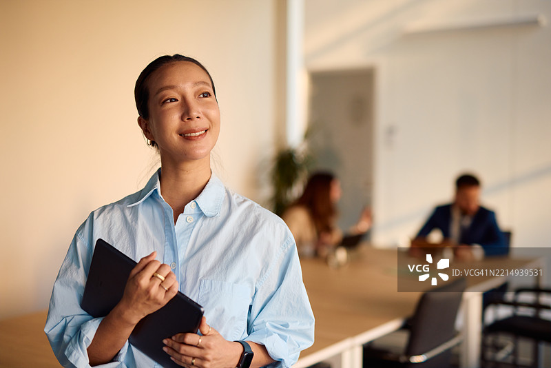 年轻的亚洲女商人在现代办公室里微笑着，手持平板电脑，目光投向一旁。图片素材