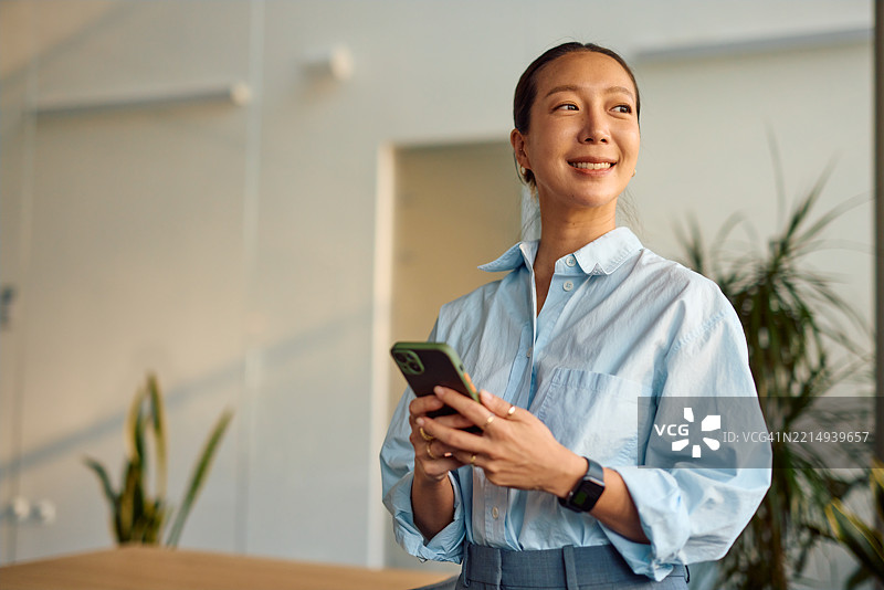 微笑的女商人在现代办公室使用智能手机并向一旁看去图片素材