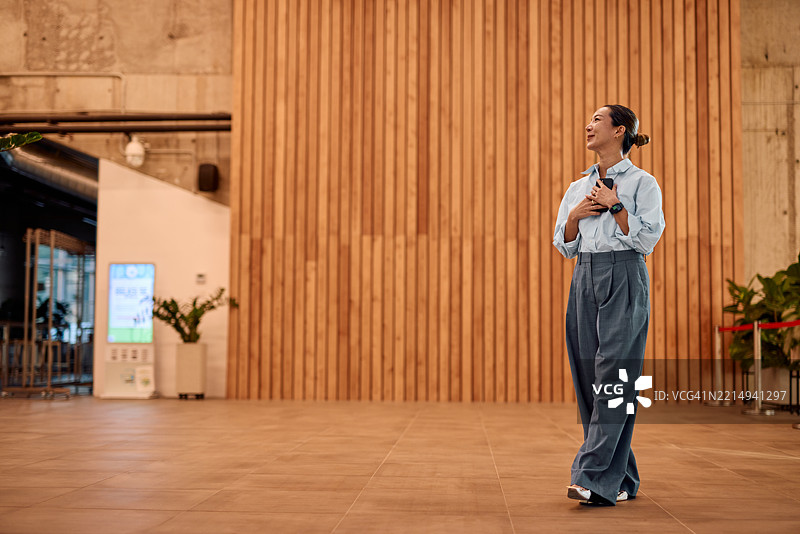 女商人在现代办公室大堂走动，手持智能手机，抬头微笑。图片素材