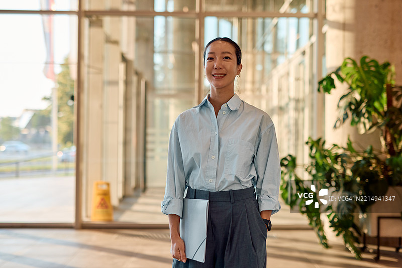 年轻的亚洲女商人在现代办公大楼中微笑并手持文件夹图片素材