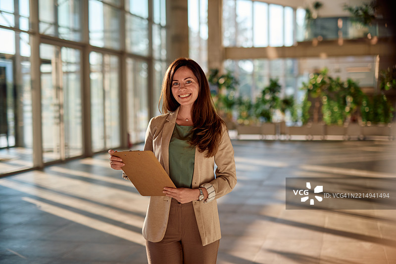 微笑的女商人在大型现代办公大楼中手持夹板图片素材