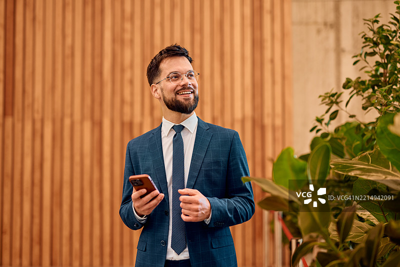 微笑的商人在现代办公室里拿着智能手机向一旁看去图片素材