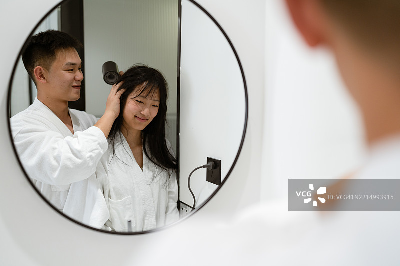 年轻男子在现代浴室镜子前为年轻女子吹干头发，愉快而亲密的时刻图片素材