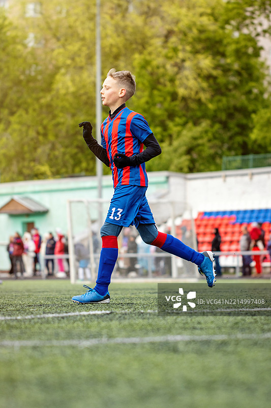 穿着13号球衣的足球运动员在场上奔跑。一名穿着蓝红条纹制服的少年在比赛中竞争，观众在旁观看。图片素材