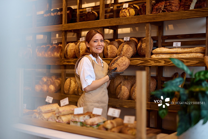 微笑的女面包师在面包店里拿着一条面包图片素材