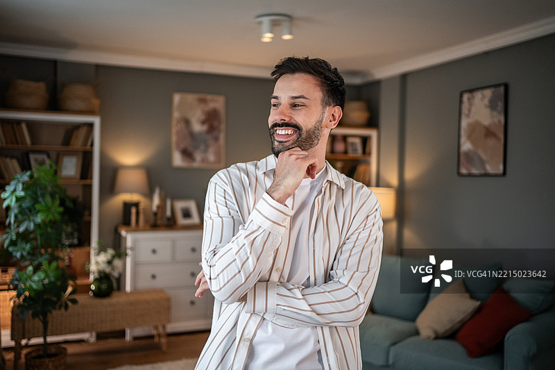 年轻男子微笑着，目光投向一旁，站在客厅里。图片素材