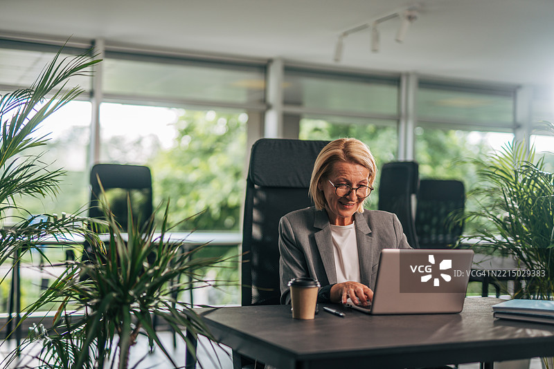 现代办公室中，身边环绕着植物的资深女商人正在使用笔记本电脑。图片素材