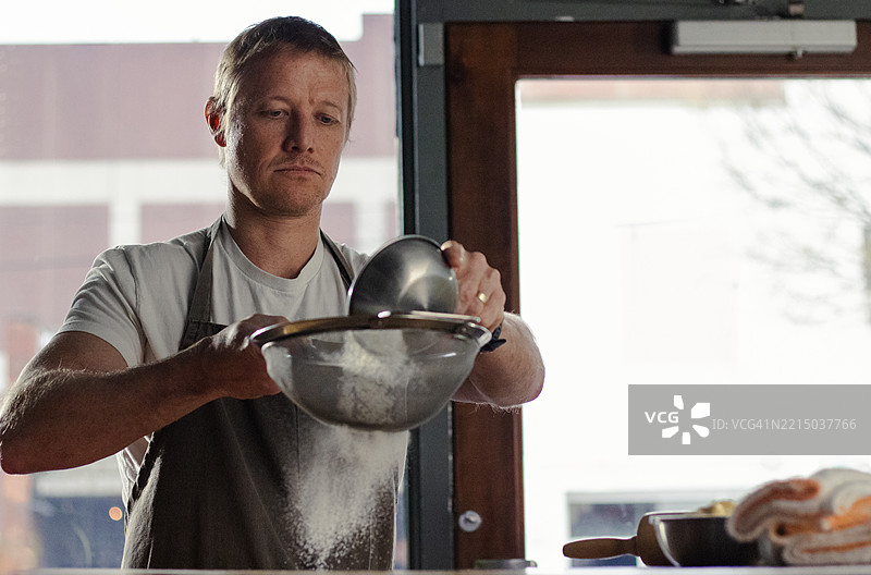 一位面包师将装满面粉的金属碗浸入筛子中，在一家光线明亮的餐厅里。图片素材