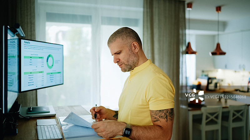 在家办公的男人在站立式办公桌前查看财务数据。图片素材