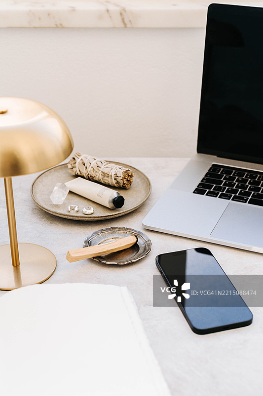 带有笔记本电脑、黑色智能手机、金色台灯、巴洛圣木、鼠尾草束和水晶的工作空间图片素材