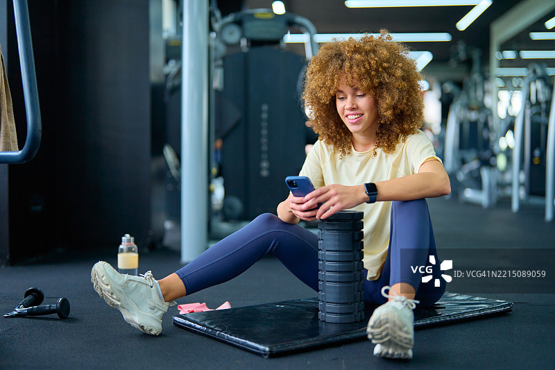 年轻女性在健身房锻炼后使用智能手机放松。图片素材