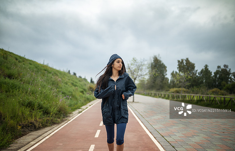 春季训练中的女性跑者在户外跑道上淋着春雨，横向视角展示一位穿着雨衣的女性在雨中跑步。图片素材