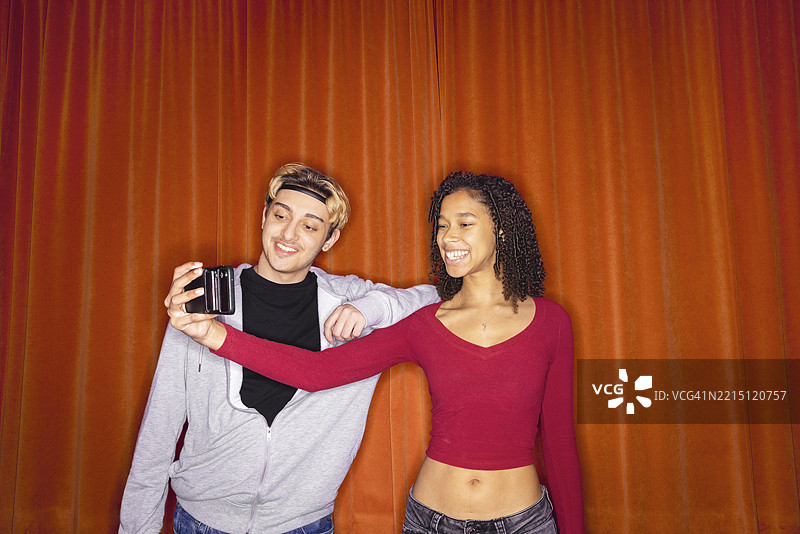 快乐的年轻女性与男性朋友在红色窗帘旁用智能手机自拍图片素材