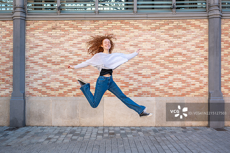 年轻的红发女性在城市背景中张开双臂跳跃图片素材