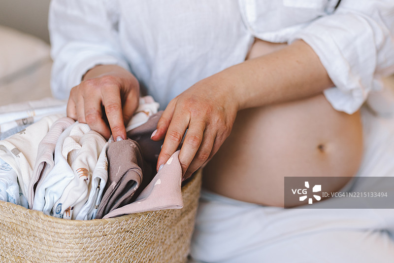 孕妇轻轻抚摸着自己的肚子，手中抱着一篮婴儿衣物。图片素材