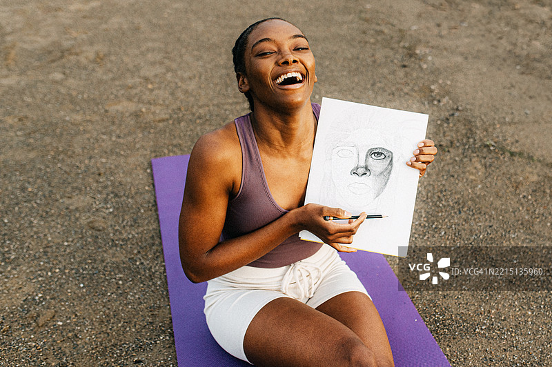 一位年轻女性坐在紫色瑜伽垫上,手里拿着一幅面孔的铅笔画,开心地笑着。图片素材