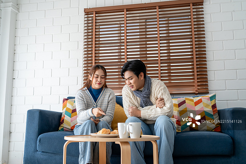 亚洲年轻情侣在冬季的家中享受温馨时光。吸引人的男士和女士穿着温暖的毛衣，感到快乐和放松，舒适地坐在家中的客厅里。图片素材