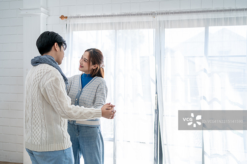 亚洲年轻夫妇在冬季的家中享受温馨时光。迷人的配偶，夫妻俩在音乐中跳舞，感到快乐和放松，舒适地站在家中的客厅里。图片素材