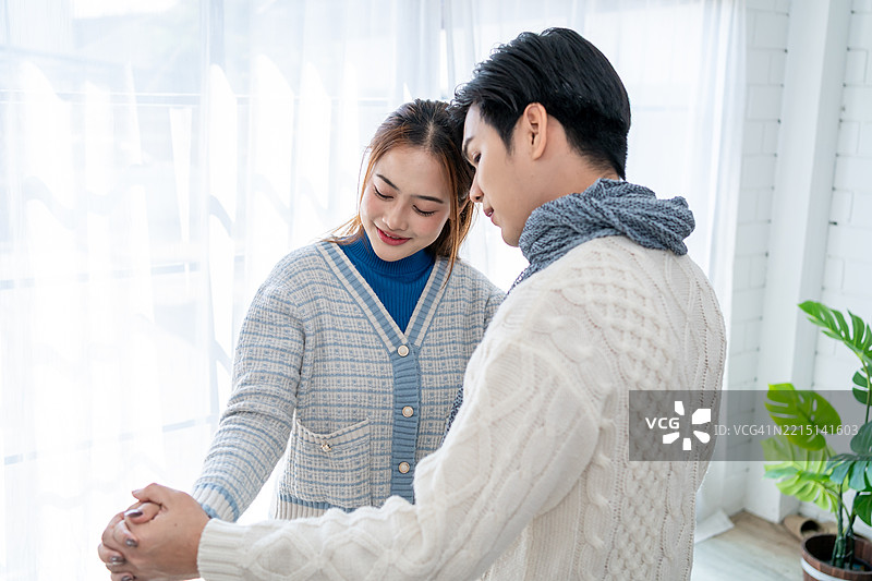 亚洲年轻夫妇在冬季的家中享受温馨时光。迷人的配偶，夫妻俩在音乐中跳舞，感到快乐和放松，舒适地站在家中的客厅里。图片素材