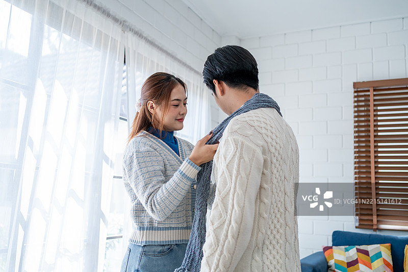 亚洲年轻夫妇在冬季的家中享受温馨时光。迷人的妻子为丈夫系上温暖的围巾，感到快乐和放松，舒适地站在家中的客厅里。图片素材