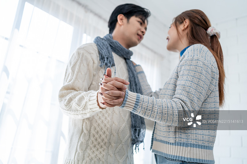 亚洲年轻夫妇在冬季的家中享受温馨时光。迷人的夫妻在客厅里舒适地站着，随着音乐跳舞，感到快乐和放松。图片素材